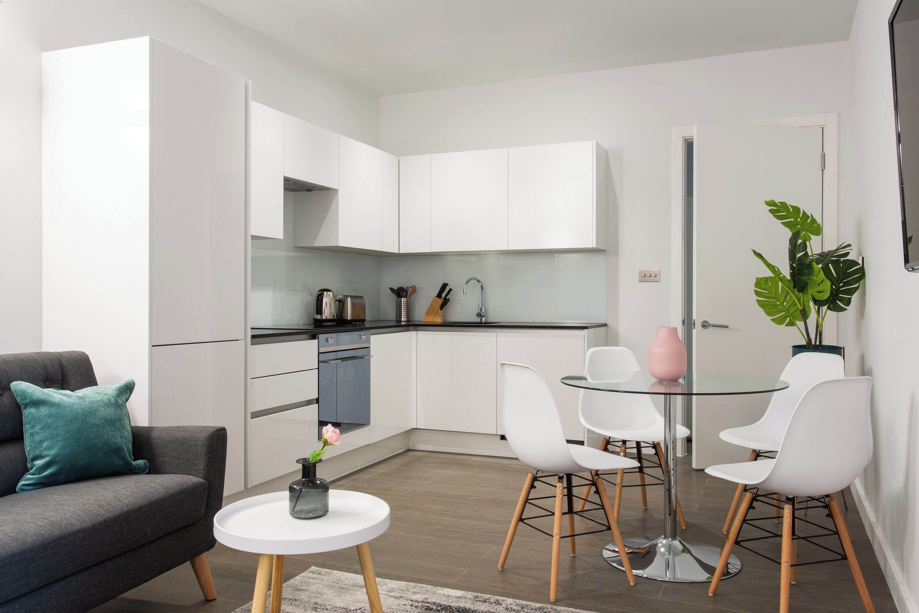A modern kitchen with white cabinets, a dining area with a glass table and white chairs, and a grey sofa with a teal pillow.