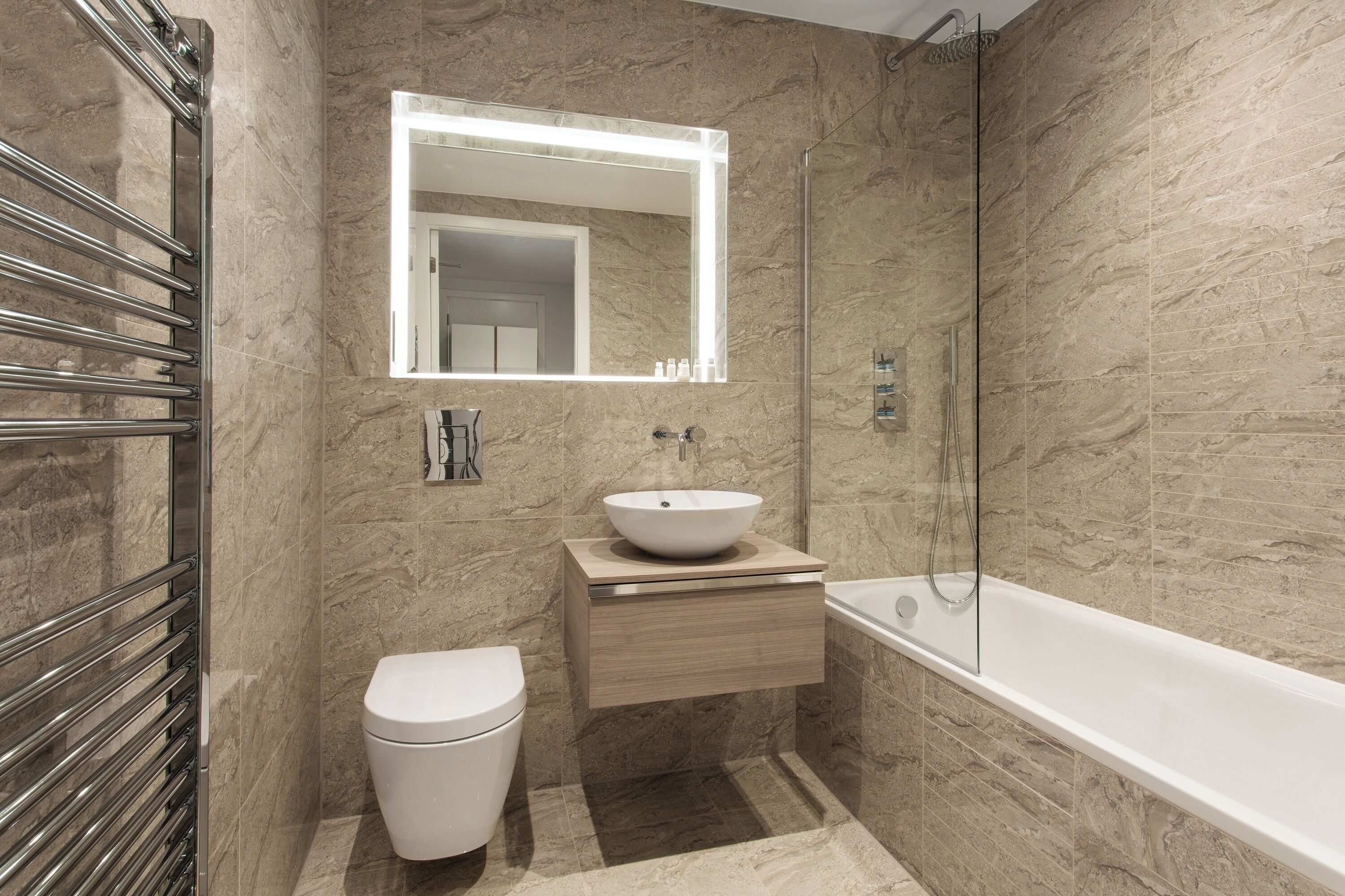 Une salle de bain moderne avec un mur en marbre, des toilettes blanches, un meuble-lavabo en bois avec un évier, un miroir avec éclairage intégré et une douche à parois de verre.
