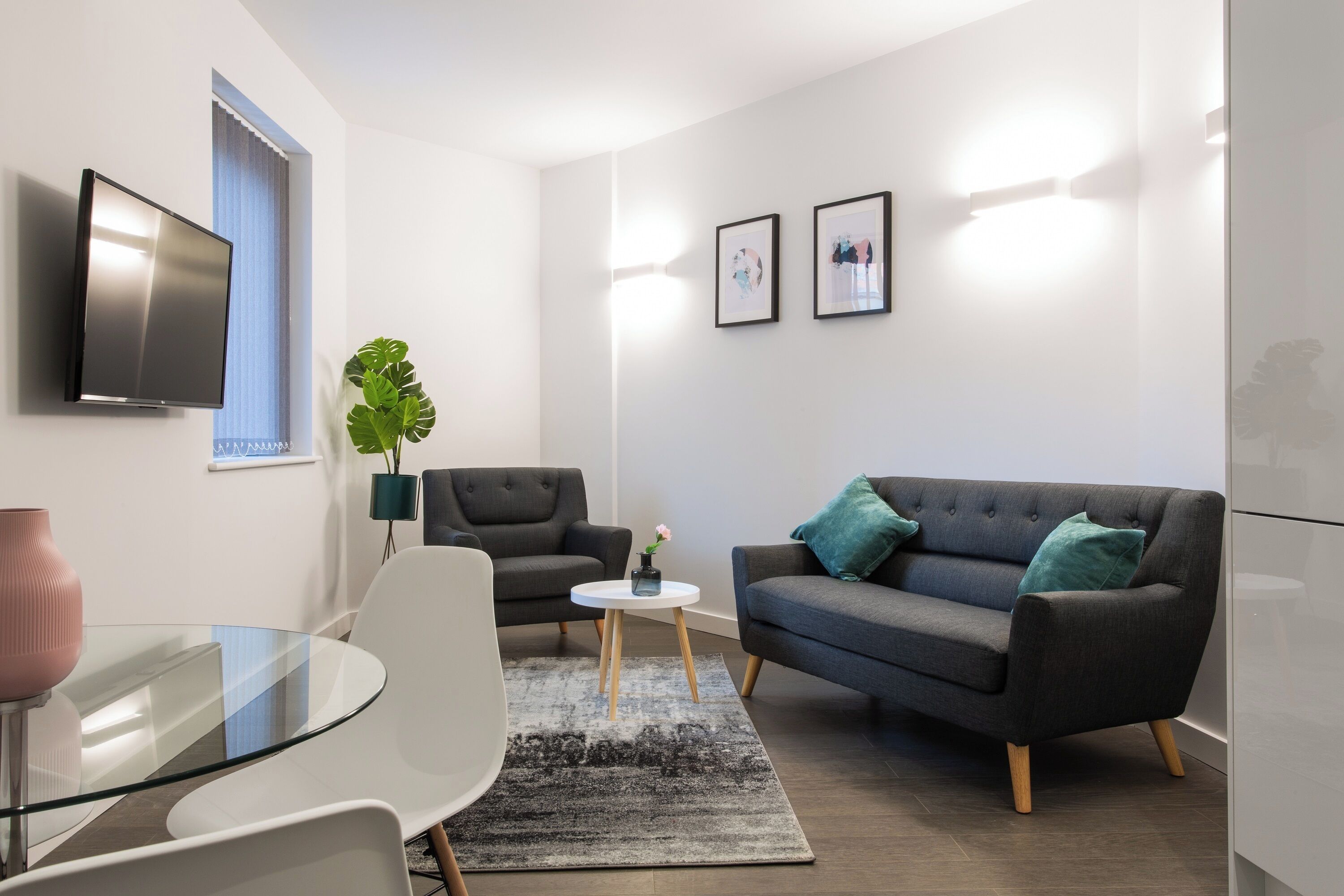 A modern living room with a glass table, two armchairs, a sofa, a TV, and framed pictures on the wall.