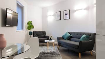 A modern living room with a glass table, two armchairs, a sofa, a TV, and framed pictures on the wall.