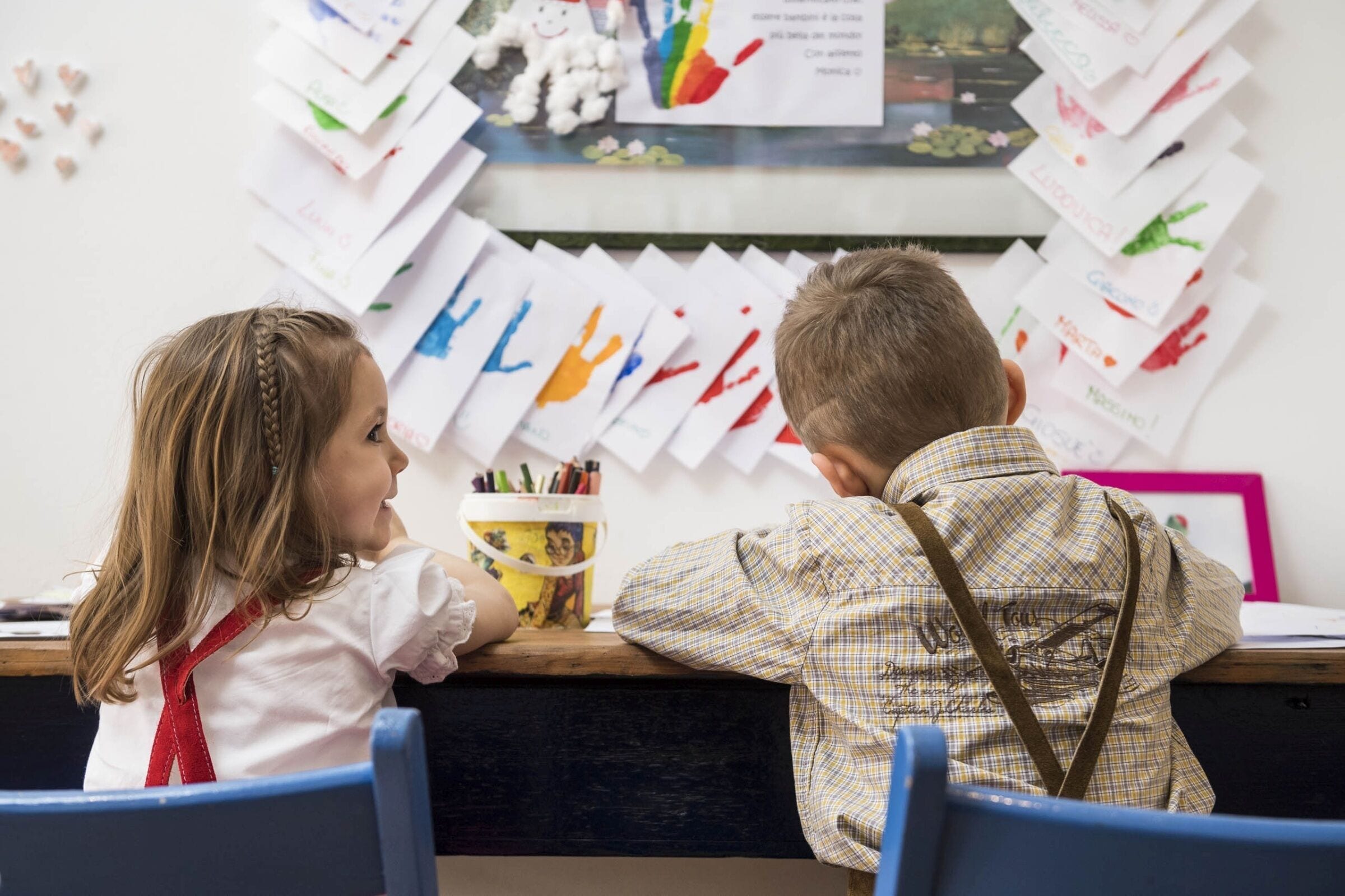 Due bambini seduti a una scrivania con fogli e un barattolo di pennarelli.