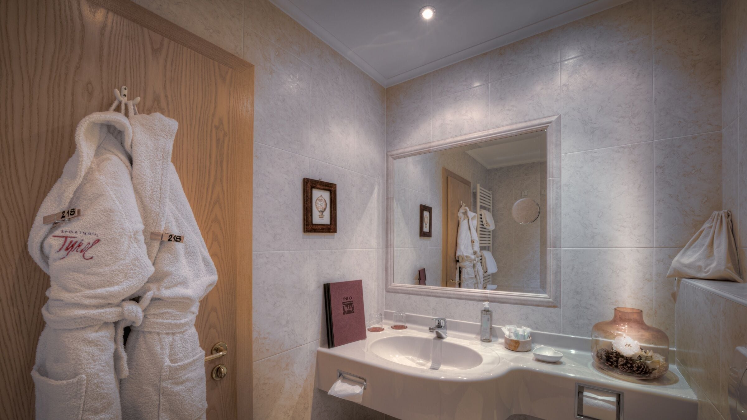 Un baño con puerta de madera, un espejo grande, un lavamanos con artículos de aseo y dos toallas blancas colgadas de ganchos.