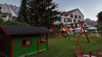 Un’area giochi con altalene e scivoli, una piccola casetta verde e, sullo sfondo, un edificio con molte finestre e balconi.