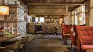 Een bar met een houten plafond, een betegelde muur met decoratieve patronen, en een bargedeelte met verschillende flessen en glazen. Op de voorgrond staan twee krukken en een tafel met stoelen.
