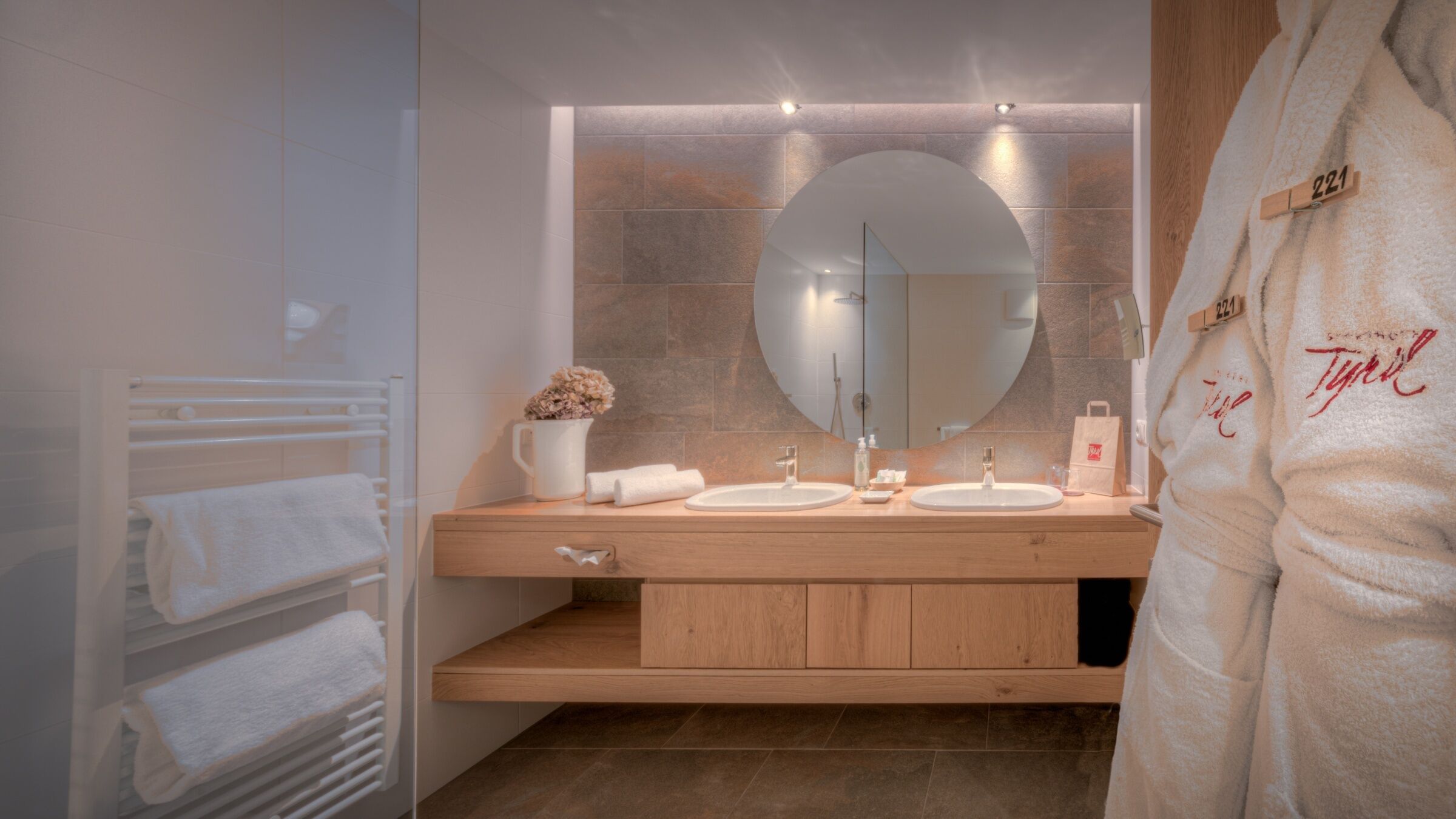 Un baño moderno con tocador de madera, dos lavamanos, un espejo grande y un perchero para toallas.