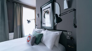 A neatly made bed with white linens and multiple pillows, a wall-mounted lamp, a framed artwork, and a window with curtains. -  The Warwick by Nestor (London)