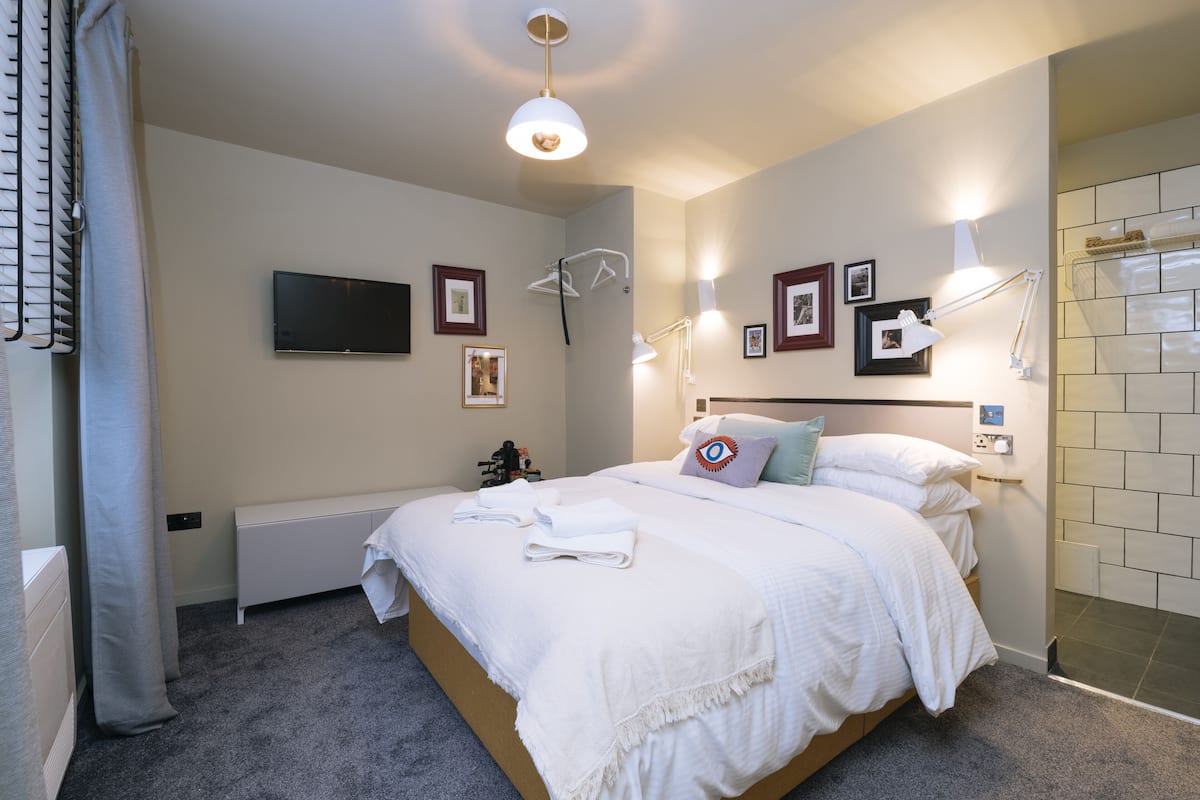 A neatly made bed with white linens, a mounted TV on the wall, and a bedside table with a lamp.