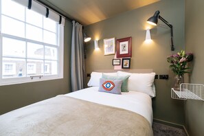 A neatly made bed with a beige blanket, a pillow with an eye design, and a vase with flowers on a nightstand. -  The Warwick by Nestor (London)