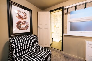 A room with a patterned armchair, a framed abstract painting, a door leading to a bathroom, and a window with blinds. -  The Warwick by Nestor (London)