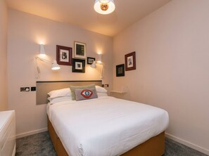 A neatly made bed with white linens, a headboard, and a pillow with an eye design. There are wall-mounted lights and framed pictures on the wall. -  The Warwick by Nestor (London)