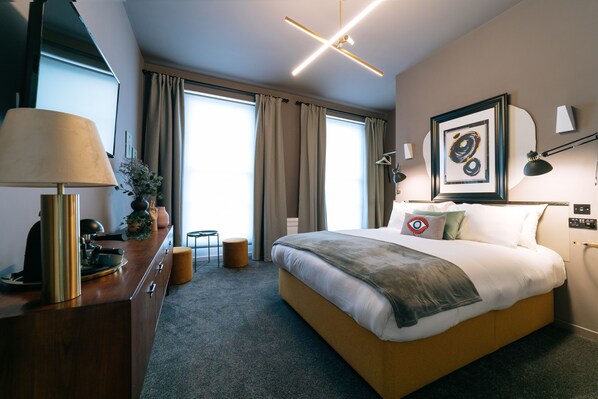 A modern hotel room with a large bed, a desk with a lamp, a framed artwork on the wall, and a view of the outdoors through the window. -  The Warwick by Nestor (London)