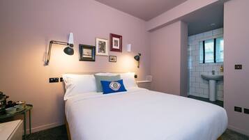 A hotel room with a bed, bedside tables, a wall-mounted lamp, framed pictures, and a bathroom with a sink and window.