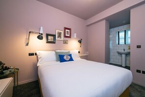 A hotel room with a bed, bedside tables, a wall-mounted lamp, framed pictures, and a bathroom with a sink and window. -  The Warwick by Nestor (London)