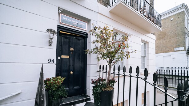 A black door with a sign above it reading "The Warwick Hotel." There is a potted plant next to the door and a black metal fence in front. -  The Warwick by Nestor (London)