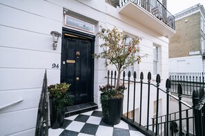A black door with a sign above it reading "The Warwick Hotel." There is a potted plant next to the door and a black metal fence in front. -  The Warwick by Nestor (London)