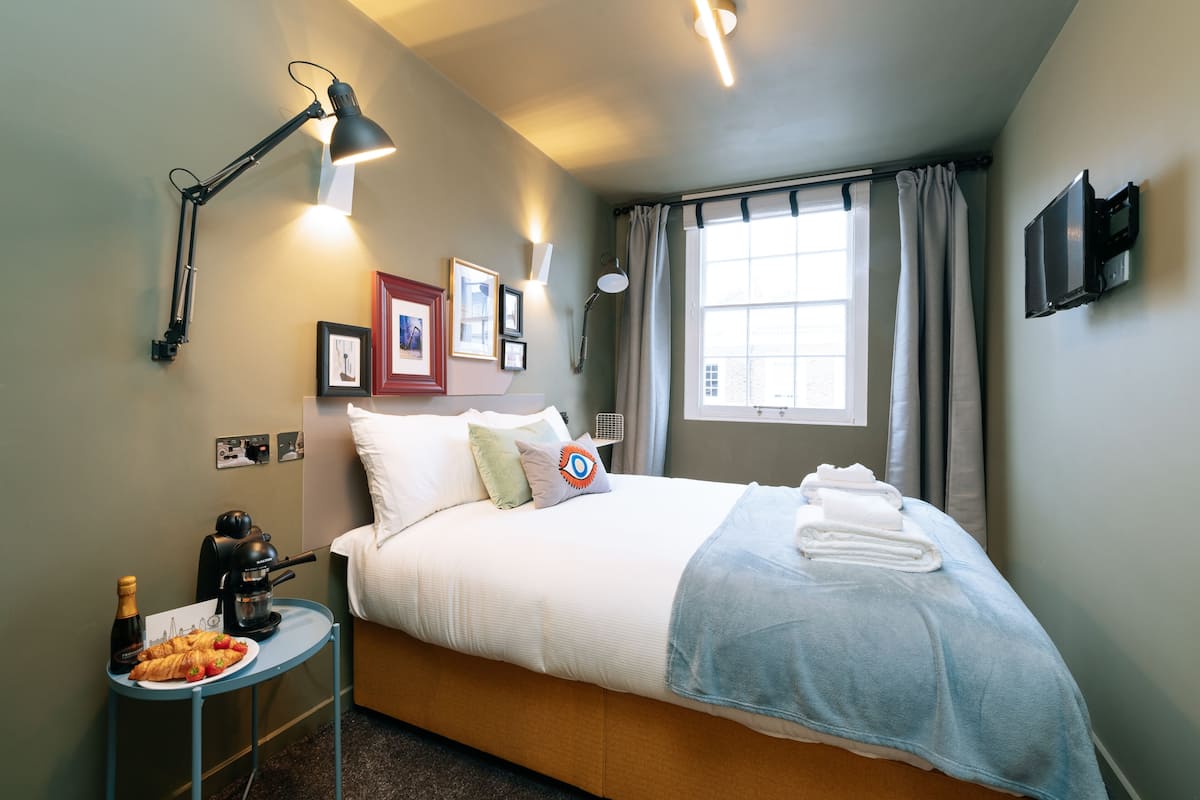 A neatly made bed with a blue blanket, a bedside table with a coffee maker and croissant, a wall-mounted TV, and a window with curtains.