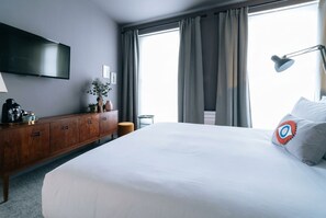 A hotel room with a large bed, a flat-screen TV mounted on the wall, a wooden dresser, a desk with a lamp, and a window with curtains. -  The Warwick by Nestor (London)