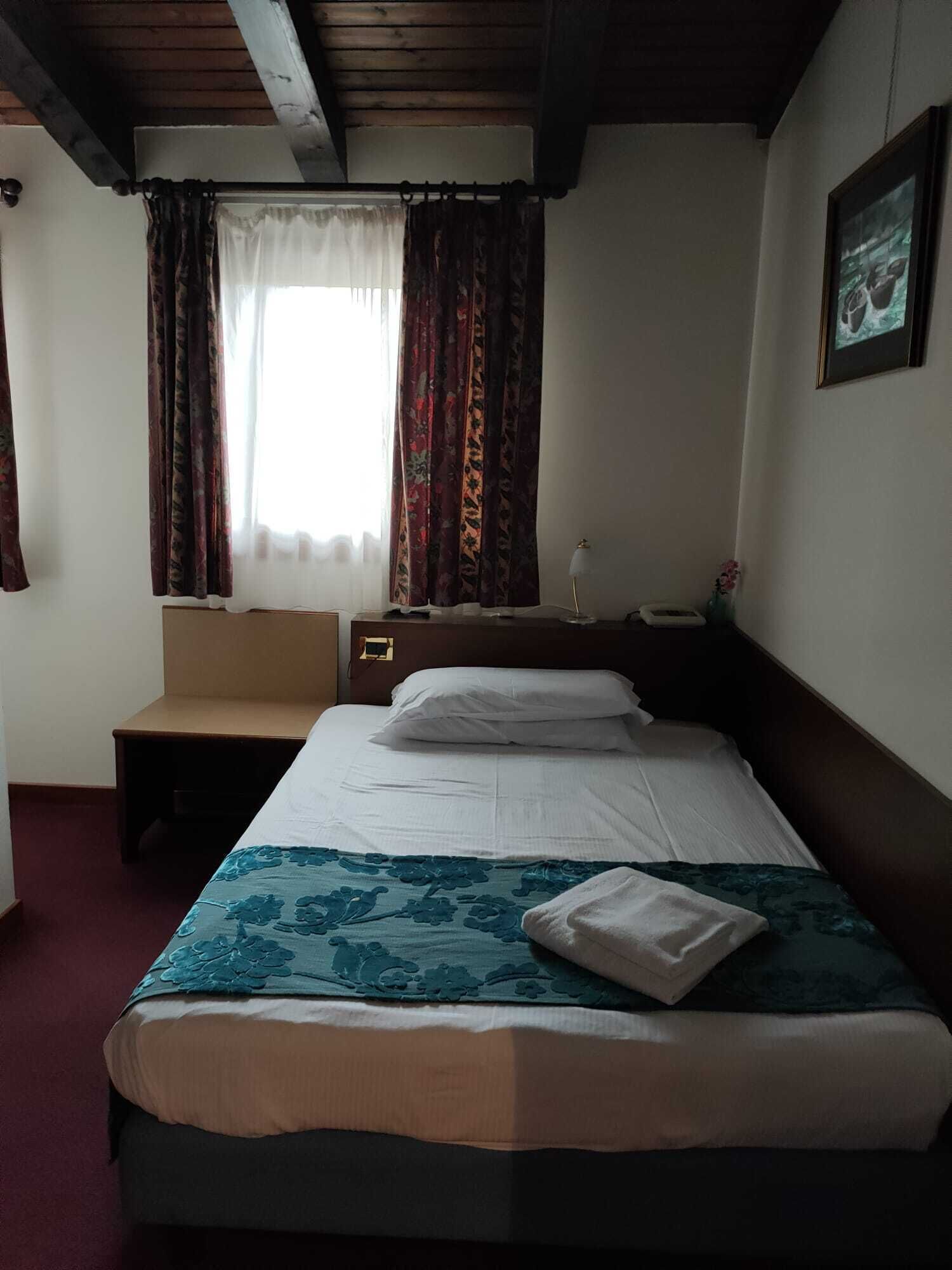 A hotel room with a bed, a bedside table, a chair, a window with curtains, and a framed picture on the wall.