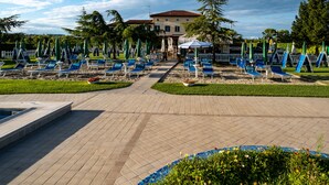 Zona de piscina con sillones reclinables y sombrillas, un camino que lleva a un edificio y un jardín ajardinado.