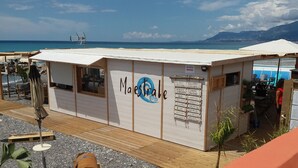 Un restaurante en la playa llamado 'Maestrale' con un cartel que enumera varios platos, mesas al aire libre con sombrillas y vistas al mar y montañas al fondo.