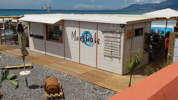 Ein Restaurant direkt am Strand namens 'Maestrale' mit einer Schautafel, die verschiedene Gerichte auflistet, Außenbereich mit Sonnenschirmen und Blick auf Meer und Berge im Hintergrund.