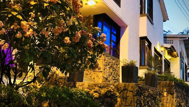 Una casa moderna de dos pisos con un muro de contención de piedra, un balcón con plantas en macetas y una entrada bien iluminada.