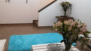 Una sala de estar con un televisor de pantalla plana montado en la pared, una escalera que sube a otro nivel, un tapete azul y un florero con flores sobre una mesa blanca.