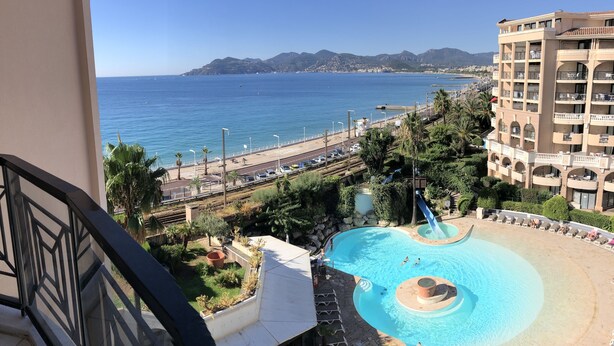 A pool area with a view of the sea and mountains, surrounded by greenery and a building with balconies. - Cannes holiday apartment 606 with balcony, sea view, A/C and parking (Provence-Alpes-Côte-D’Azur)