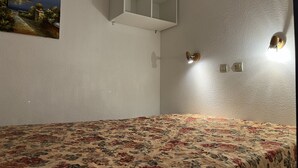 A bedroom with a floral bedspread, a painting of a coastal scene, and a built-in shelf with a light fixture. - Cannes holiday apartment 606 with balcony, sea view, A/C and parking (Provence-Alpes-Côte-D’Azur)
