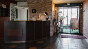 A reception area with a brick wall, a wooden reception desk, a glass door leading outside, and a tiled floor. - Le Mas De Gaujac (Lezignan-Corbieres)