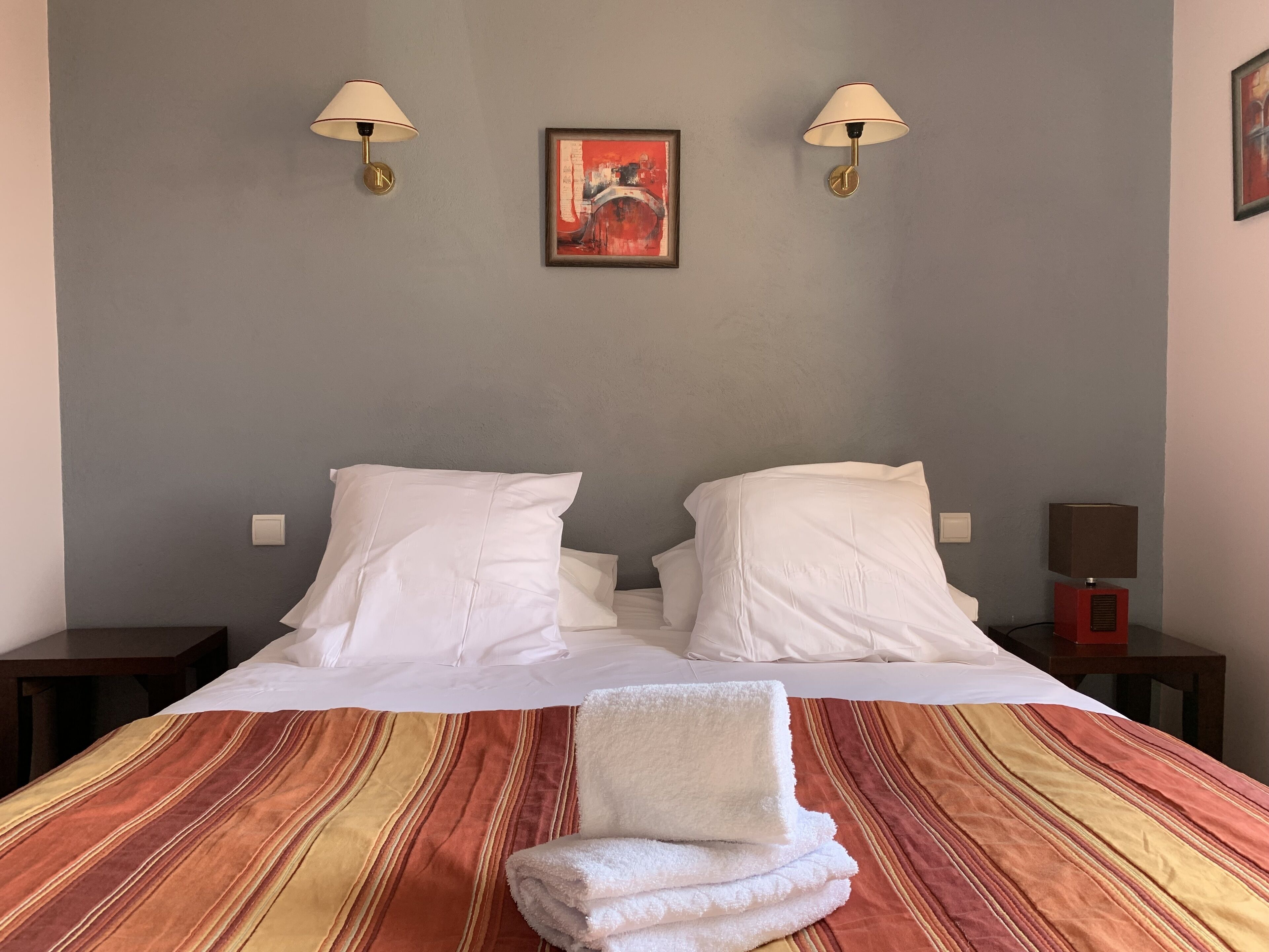 Habitación de hotel con cama, dos lámparas de noche, un cuadro en la pared y una colcha de rayas doradas y rojas.