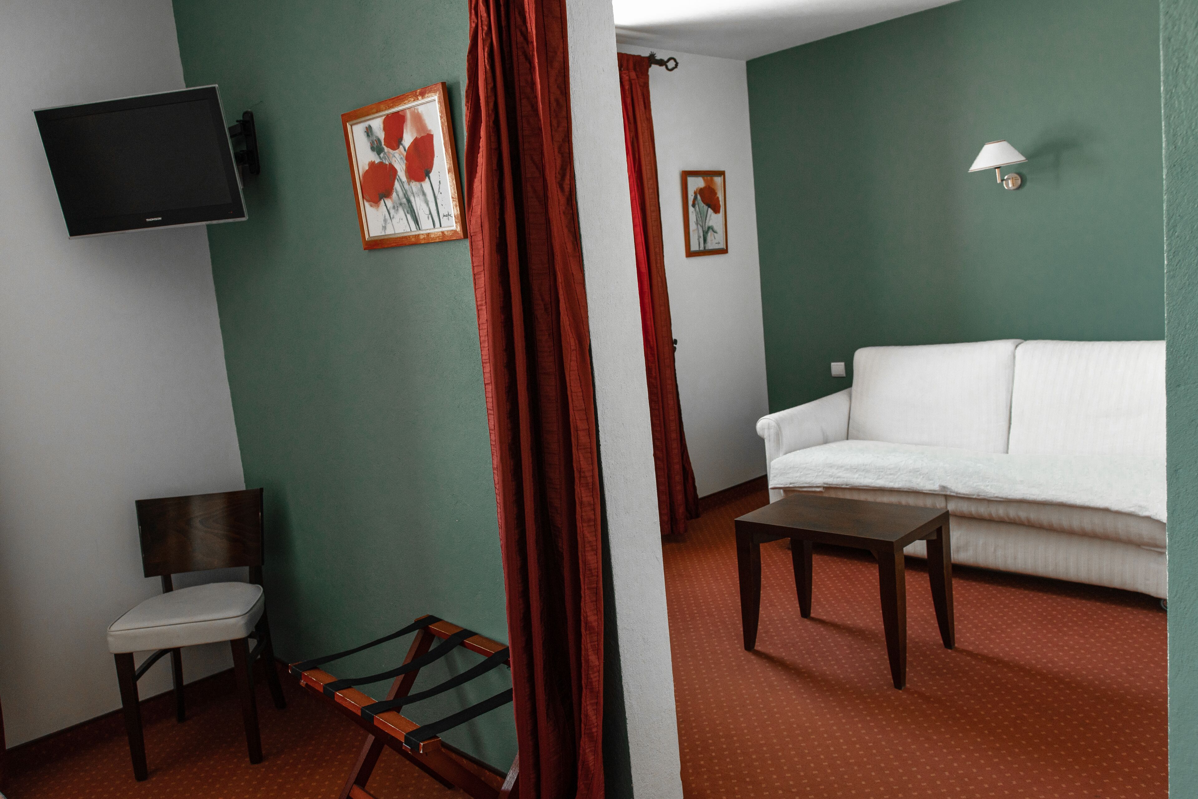 Una habitación con un sofá blanco, una silla de madera, una mesita pequeña, un televisor montado en la pared y dos cuadros enmarcados.