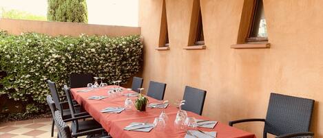 A dining area with a long table set for six, surrounded by wicker chairs, under a pergola with a tiled floor.