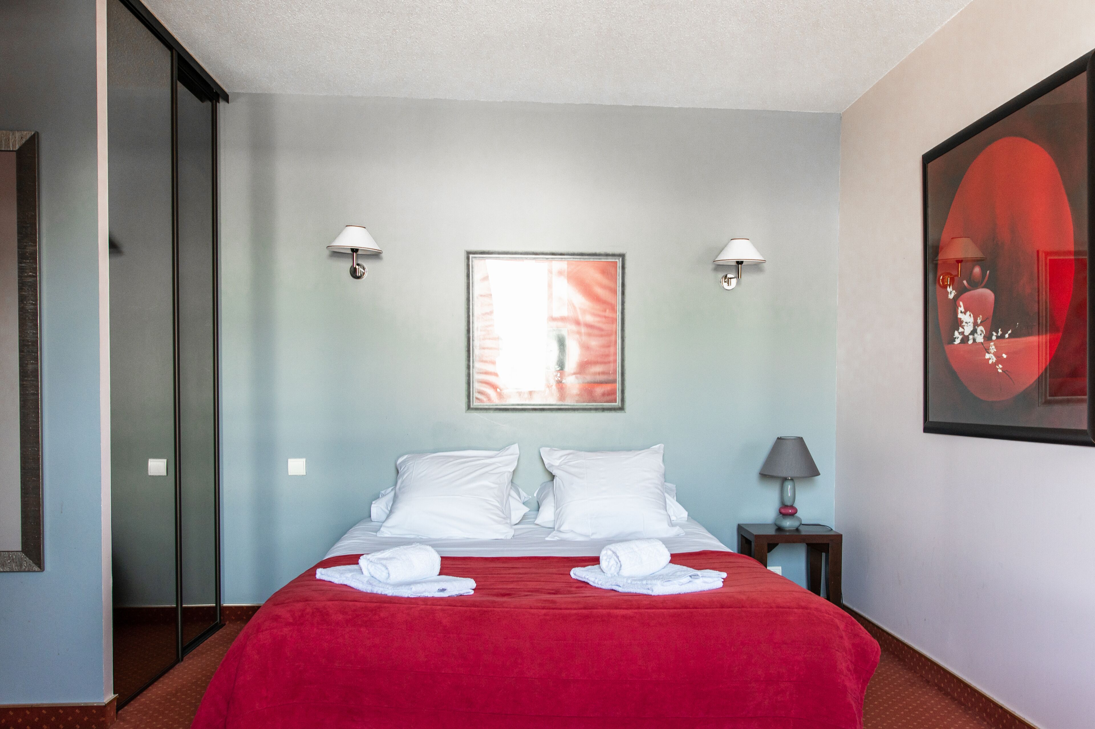 Habitació d'hotel amb un llit, dues làmpades penjades a la paret, un quadre amb marc i una manta vermella.