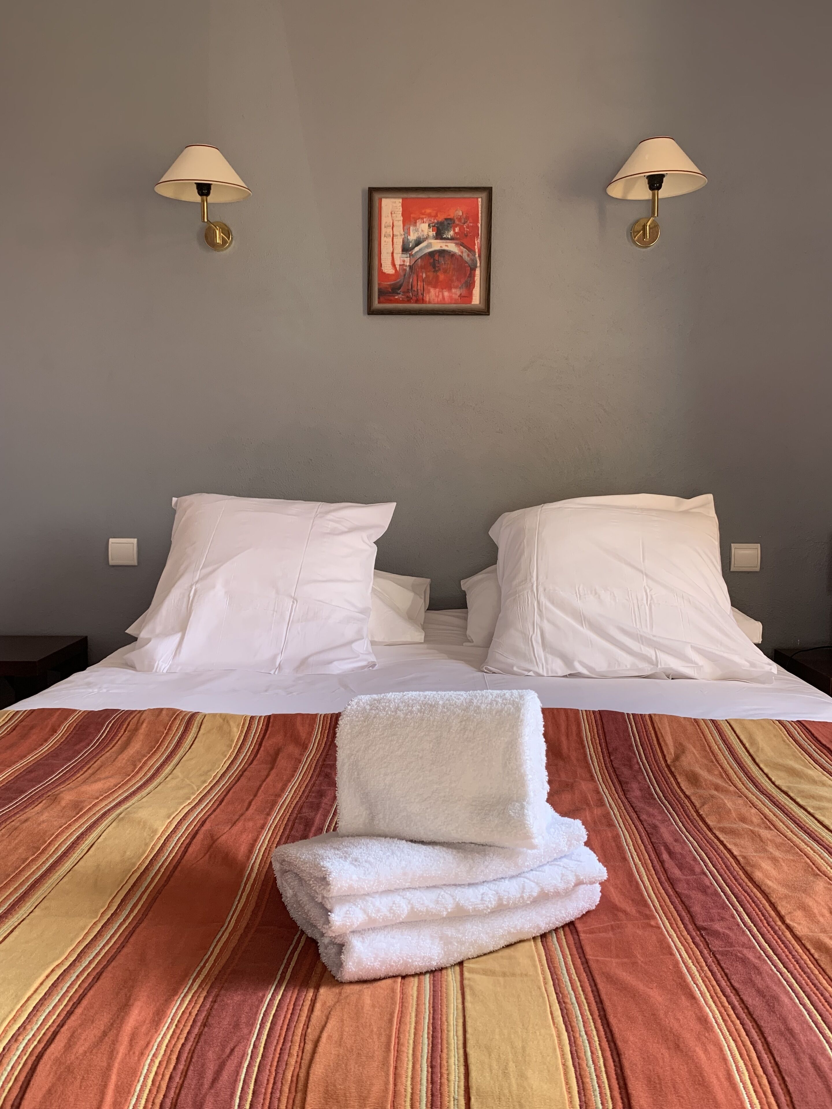 Un lit double avec du linge de lit blanc, une couvre-lit à rayures et une serviette pliée posée sur le lit. Deux lampes murales et un tableau encadré sont accrochés au mur, au-dessus du lit.