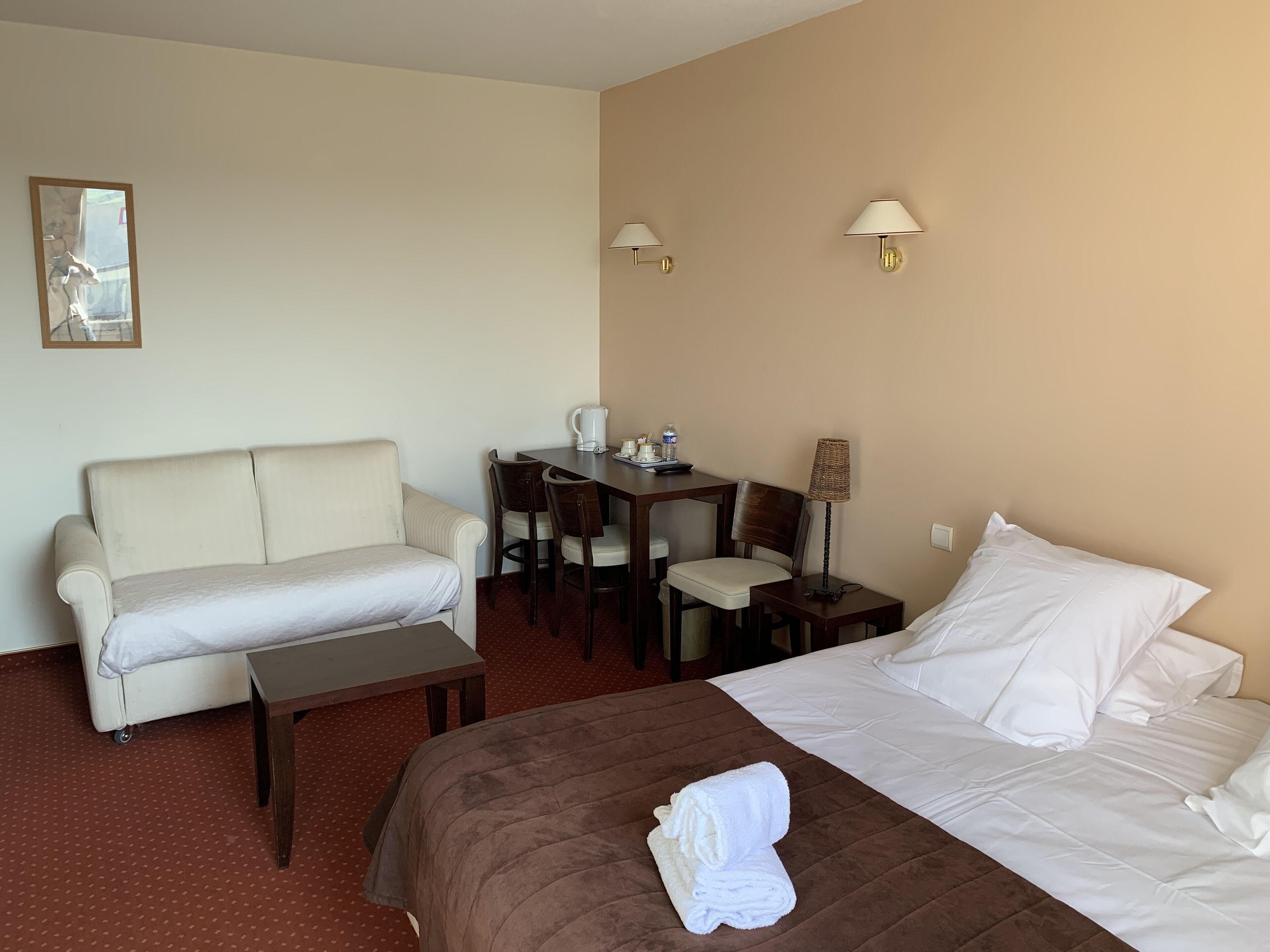 Quarto de hotel com cama, sofá, mesinha, escrivaninha com cadeiras e duas luminárias de parede.