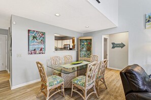 A dining area with a glass table and wicker chairs, a kitchen in the background, and a 'BEACH' sign on the wall.