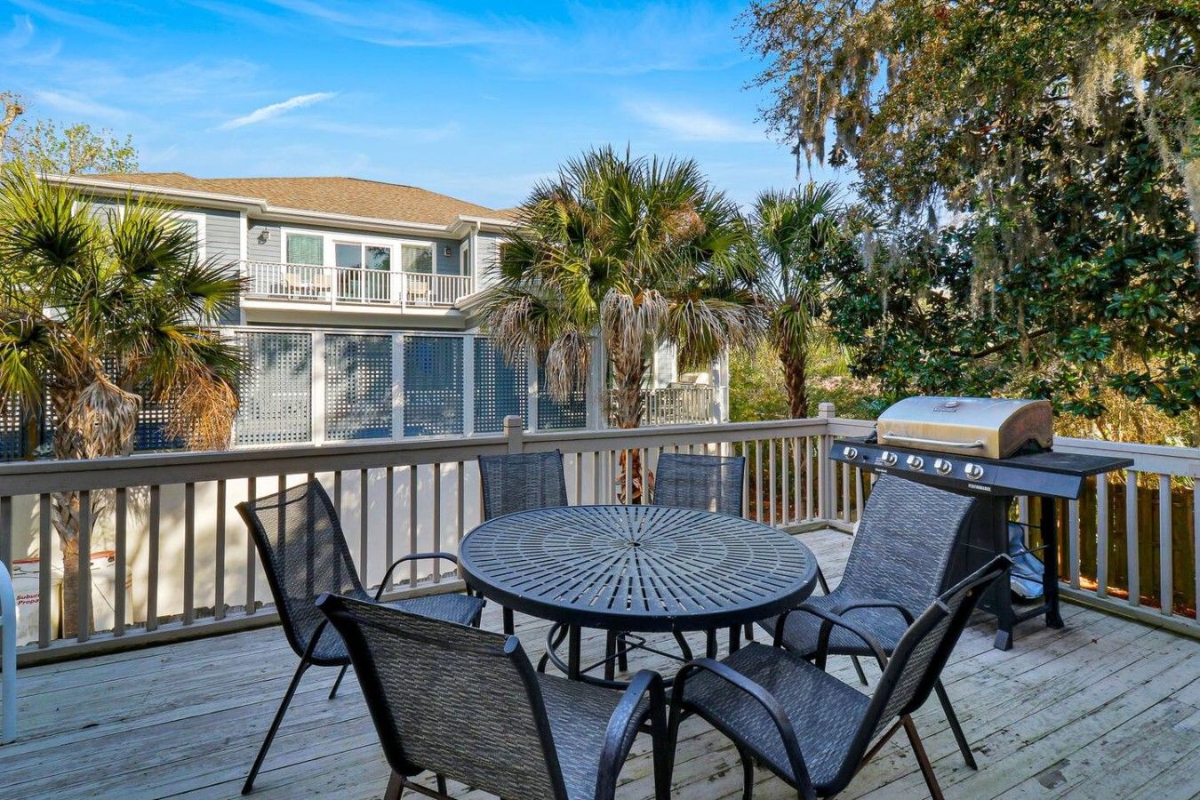 A deck with a round table and chairs, a grill, and a view of a house.