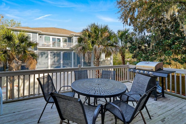 A deck with a round table and chairs, a grill, and a view of a house.