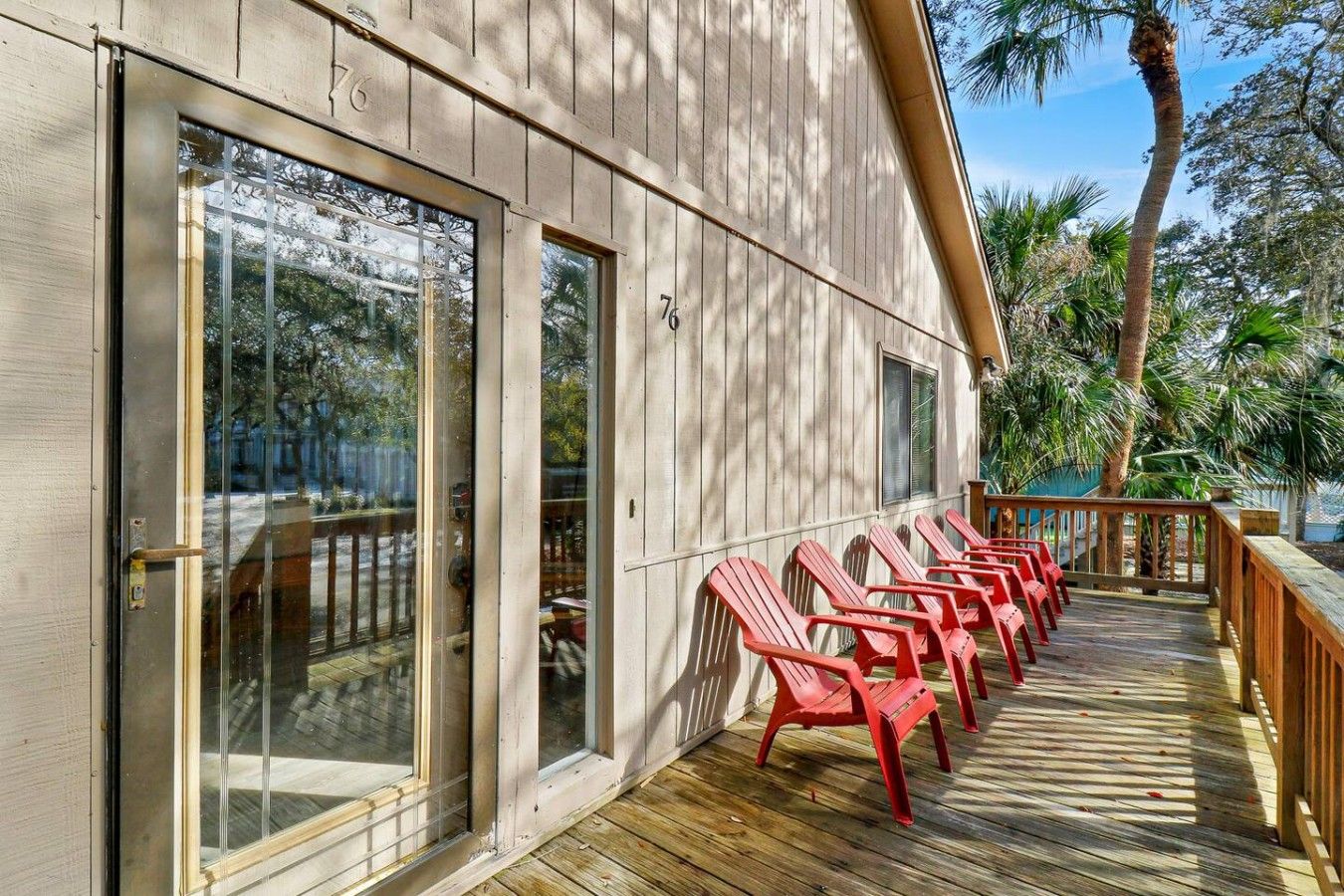 Une terrasse en bois avec des chaises rouges, une porte d’entrée en verre et une vue sur des arbres et un palmier.