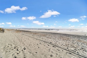 A beach with a sandy path leading to the water, a wooden fence on the left, and a clear sky with clouds.