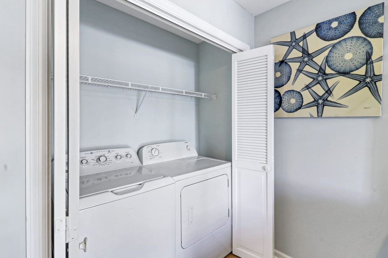 A laundry room with a white washer and dryer, a drying rack, and a wall-mounted artwork.