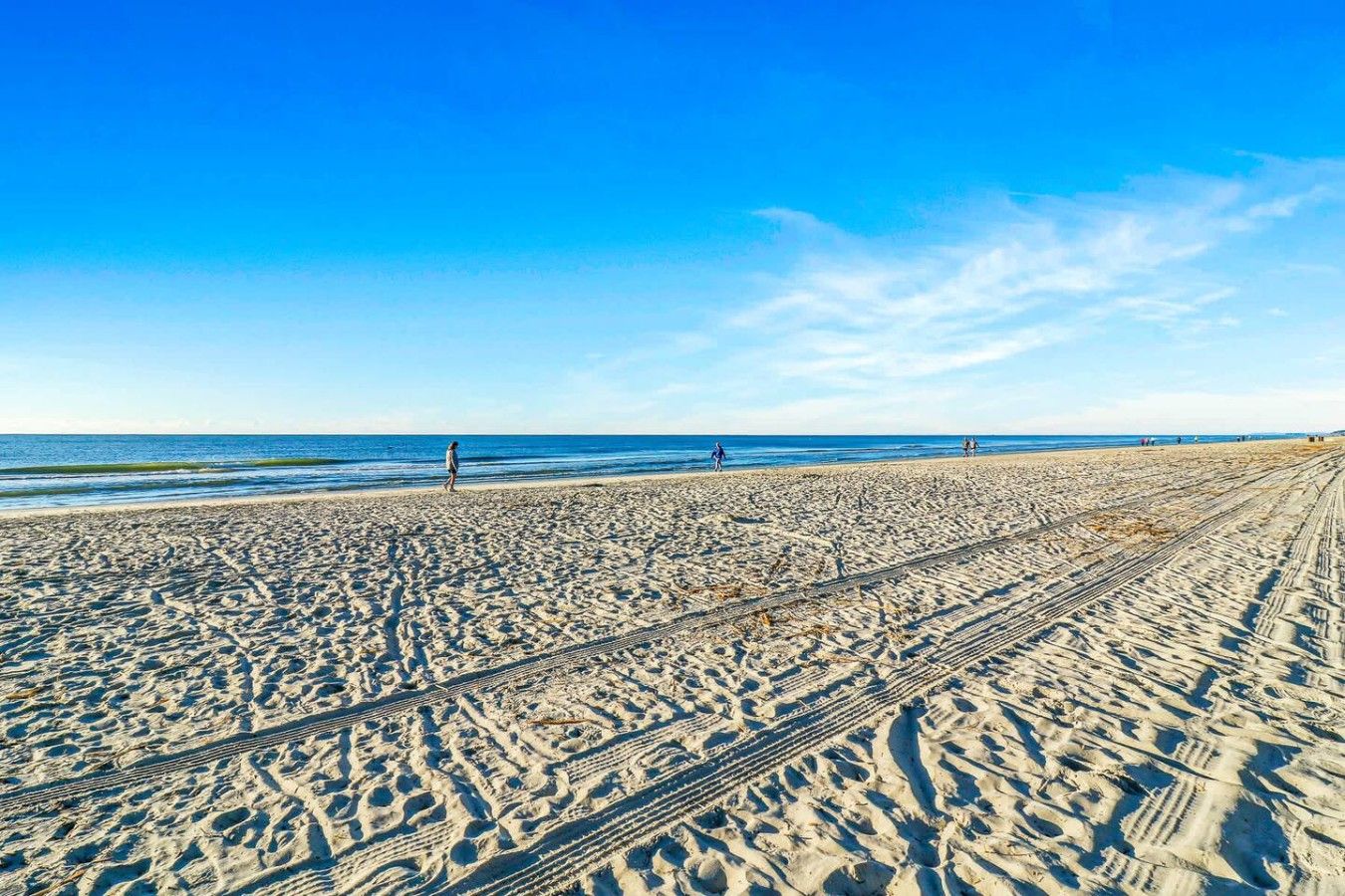 Une plage de sable avec des traces de pneus, un ciel bleu dégagé et l’océan en arrière-plan.