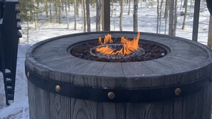 A round, wooden outdoor fire pit with metal accents, surrounded by a wooden deck and snow-covered ground, with a forest in the background. - Snowmobile, Skiing, and Outdoor Paradise/Cabin (Niagara)