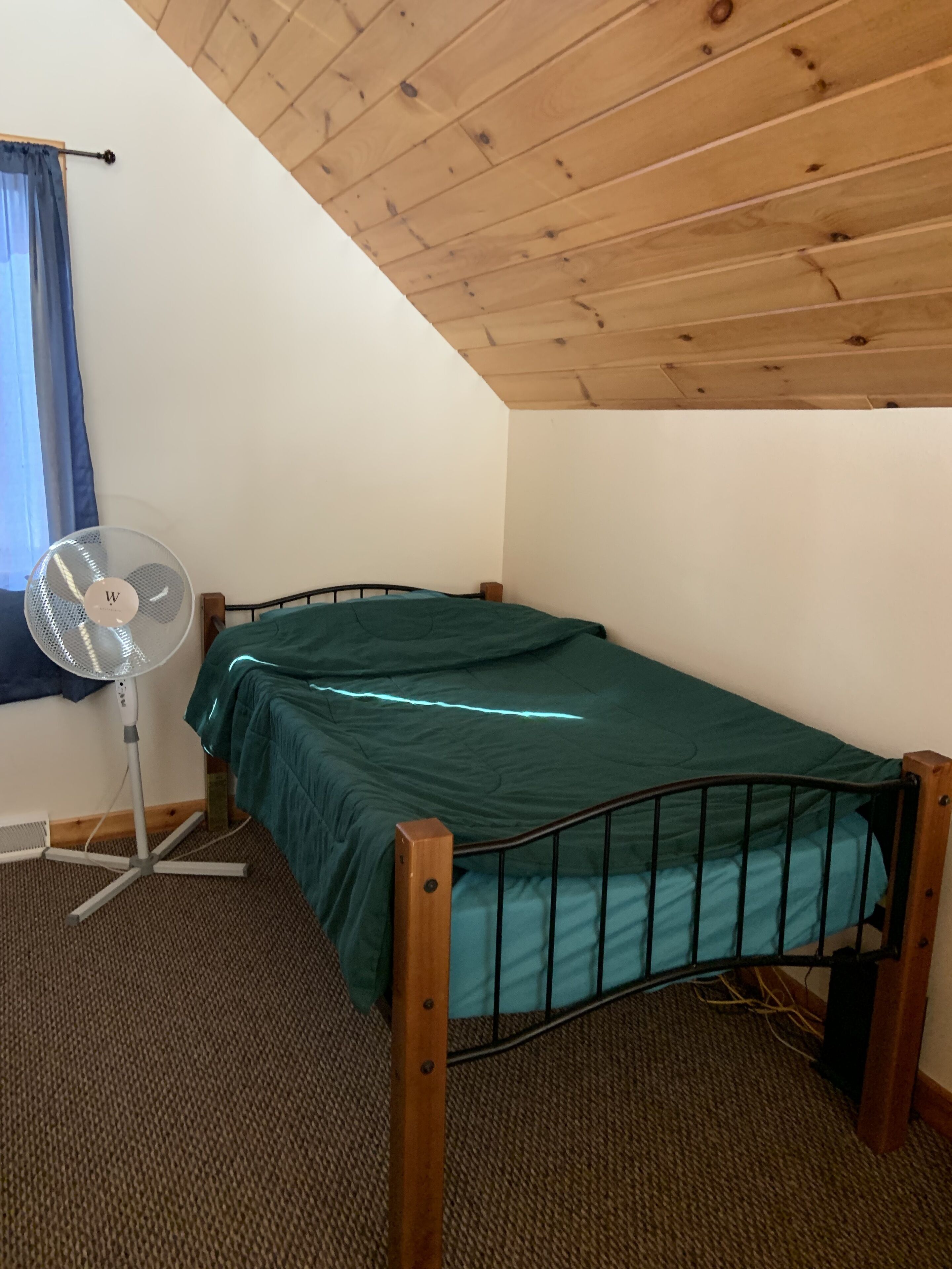 Ein Einzelbett mit grüner Matratze in einem Zimmer, das sichtbare Holzbalken an der Decke, einen Standventilator und ein Fenster mit blauen Vorhängen hat.