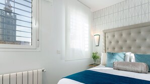 Une chambre avec une grande fenêtre, un radiateur, un lit recouvert d’une couvre-lit bleue et blanche, et un luminaire fixé au mur.