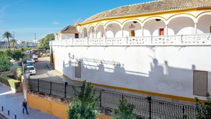 Un bâtiment blanc avec un toit en tuiles et des arcades, une rue pavée avec des voitures, et un piéton.