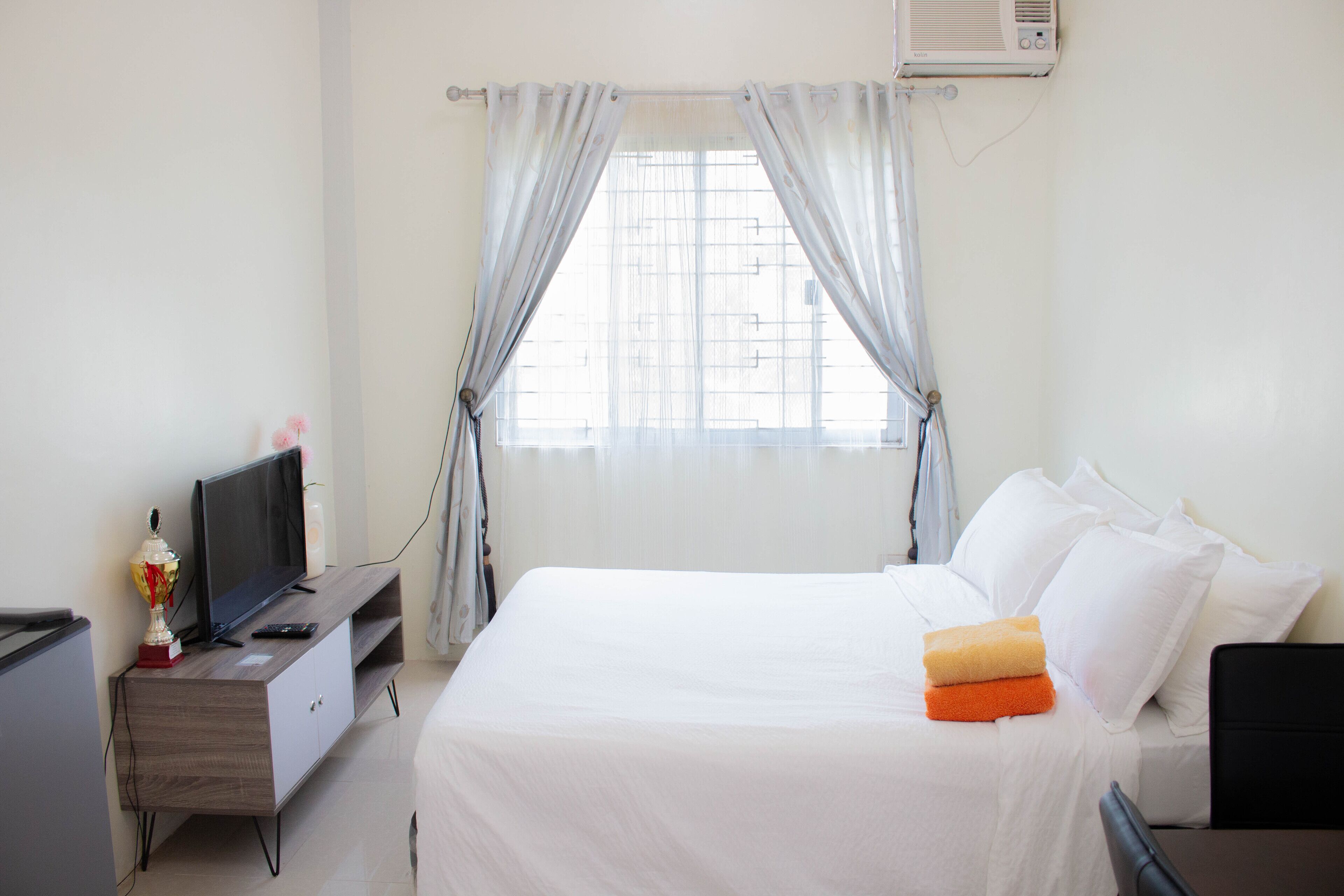 A hotel room with a bed, a TV on a wooden stand, a window with curtains, and an air conditioning unit.
