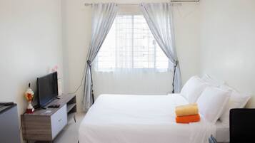 A hotel room with a bed, a TV on a wooden stand, a window with curtains, and an air conditioning unit.