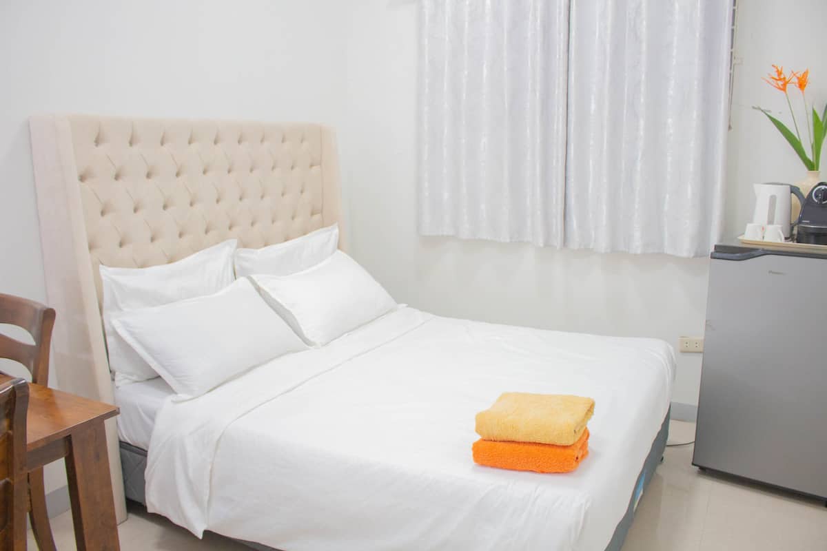 Uma cama bem arrumada com lençóis brancos, uma cadeira de madeira e uma mesa pequena com uma cafeteira.